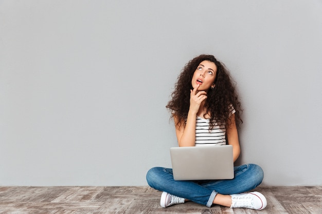 Retrato de mulher sonha em roupas casuais, sentado com as pernas cruzadas no chão com o rosto para cima, trabalhando no computador prateado sobre parede cinza