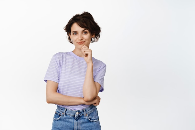 Foto grátis retrato de mulher sorridente confiante, rosto presunçoso sorridente e olhando determinada garota pensativa e criativa tem um plano, de pé contra um fundo branco. copiar espaço