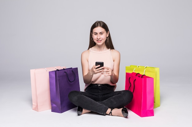 Retrato de mulher sorridente, sentado no chão com sacolas de compras e segurando o telefone móvel sobre parede cinza