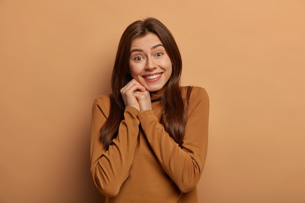 Retrato de mulher terna sorri amplamente, tem dentes brancos, gosta de momentos agradáveis, usa gola olímpica, isolado sobre o espaço marrom