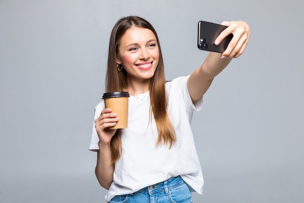Retrato de mulher tirando foto de selfie em smartphone no escritório e bebendo café para viagem em copo plástico isolado