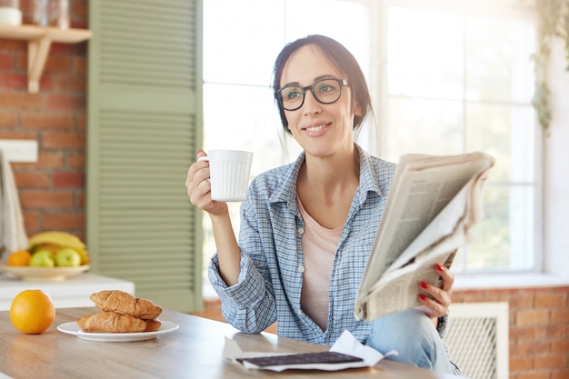 Retrato de mulher toma café da manhã, bebe café ou chá com croissants e chocolate, lê jornal sozinha em casa.