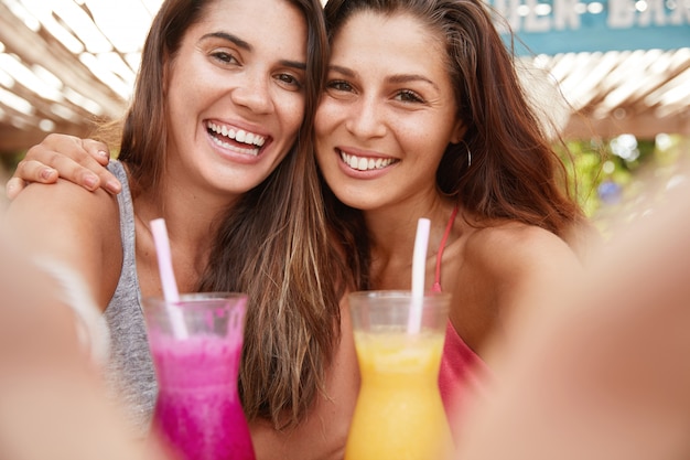 Retrato de mulheres jovens alegres com expressão alegre, segurando coquetéis e fazendo selfie, mostrando dentes brancos perfeitos e sorrisos largos
