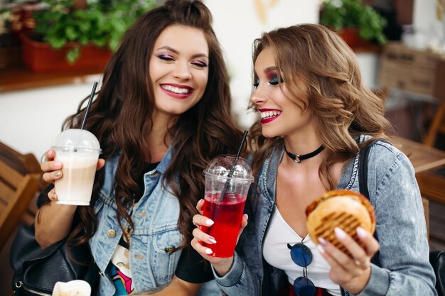 Retrato de mulheres lindas alegres desfrutando de limonada e café e hambúrgueres no café de fast food.