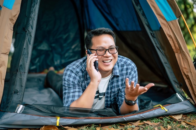 Retrato de óculos de homem viajante asiático feliz mentindo e falando ob móvel em acampamento de tendas acampamento ao ar livre e conceito de estilo de vida