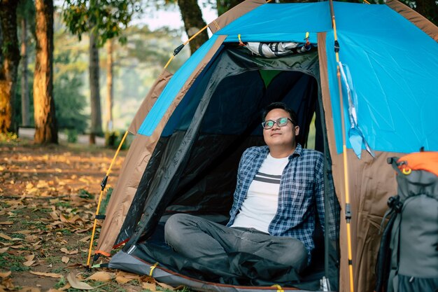 Retrato de óculos relaxantes de viajante asiático olhando para a vista enquanto está sentado em uma barraca de acampamento Acampamento ao ar livre e conceito de estilo de vida