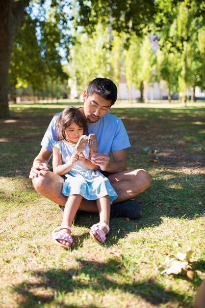 Retrato de pai asiático e sua filha com sorvete no parque. Homem feliz com a garota de joelhos, sentado na grama, ambos segurando sorvete e olhando para ele. Conceito de descanso de paternidade e verão ao ar livre