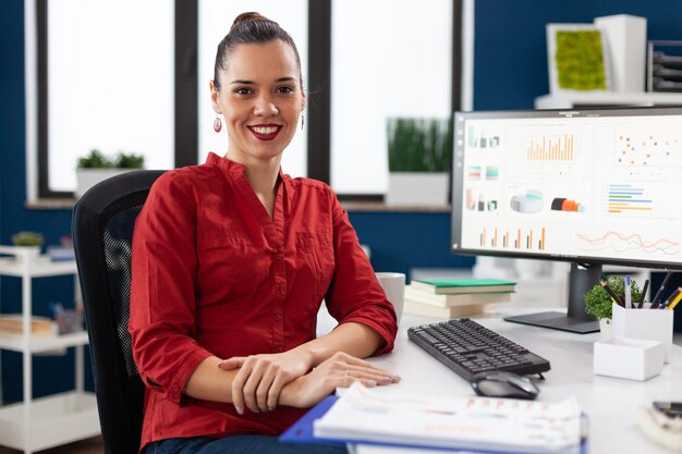 Foto grátis retrato de pequeno empresário sorridente no escritório de arranque. empresário de camisa vermelha parecendo confiante na mesa com prancheta e computador desktop. gerente posando com resultados de rotatividade.