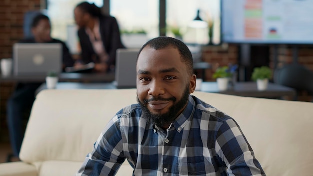 Retrato de pessoa afro-americana sorrindo no escritório, sentado no sofá para trabalhar no laptop. Funcionário da empresa usando estatísticas financeiras para criar desenvolvimento de empregos e crescimento de negócios.