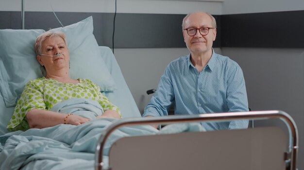 Retrato de pessoas idosas sentadas na enfermaria do hospital, a mulher mais velha com doença deitada na cama e o marido dando apoio e ajuda. Idoso ajudando pensionista com doença na clínica.