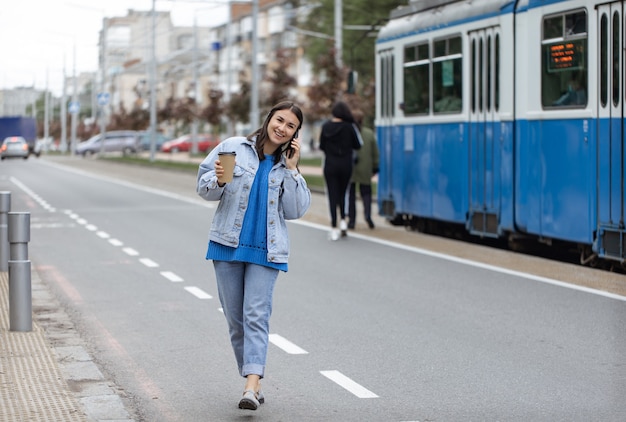 Retrato de rua de uma jovem alegre, falando ao telefone com café na mão.