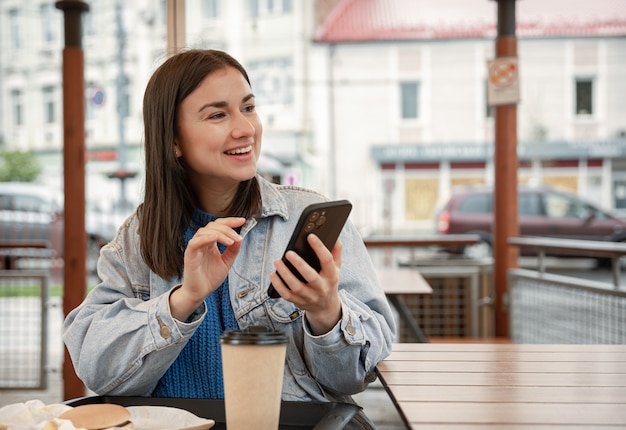 Retrato de rua de uma jovem alegre no terraço de um café, segurando um telefone.