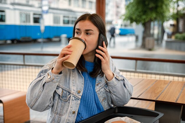 Retrato de rua de uma jovem que está tomando café, falando ao telefone e esperando por alguém.