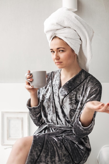 Retrato de senhora de roupão de veludo sentado com toalha na cabeça e copo na mão enquanto pensativamente olhando para a câmera em casa