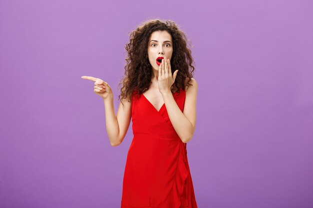 Retrato de senhora elegante encantadora surpresa e espantada com maquiagem de noite em vestido vermelho cobrindo a boca aberta de choque e espanto apontando para a esquerda curiosa e questionada sobre fundo roxo.