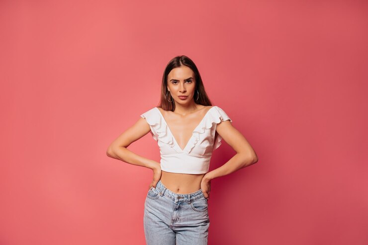 Foto grátis retrato de senhora morena elegante com top branco olhando para a frente com os braços na cintura, fazendo um grande sorriso isolado na parede rosa brilhante