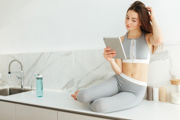 Retrato de senhora pensativa em top esportivo e leggings sentado na mesa da cozinha com tablet na mão