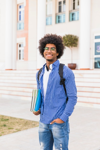 Retrato, de, sorrindo, jovem, macho afro, estudante, segurando, livros, em, mão, ficar, contra, universidade, predios