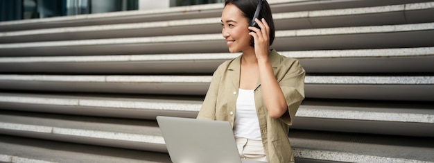 Retrato de sorrisos elegantes de uma jovem sentada nas escadas com fones de ouvido e laptop trabalhando na lição de casa