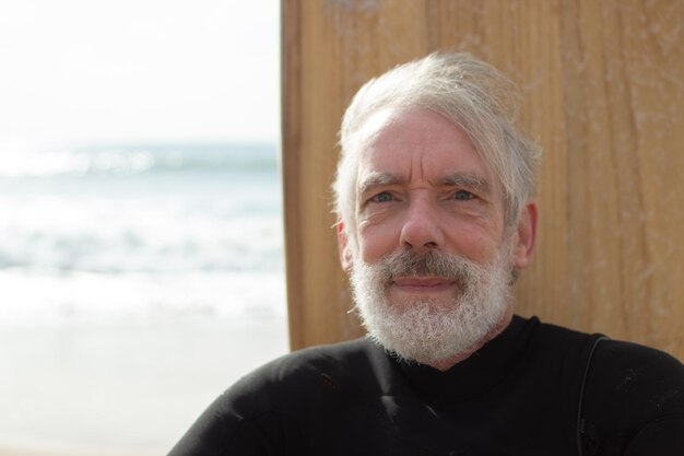 Retrato de surfista masculino envelhecido pensativo. Homem barbudo sentado na costa arenosa com placa em pé atrás das costas, olhando para a câmera no fundo do mar. Estilo de vida saudável para o conceito de pessoas idosas