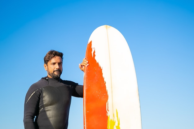 Retrato de surfista pensativo em pé com a prancha. Homem caucasiano, morena, vestindo roupa de mergulho, segurando uma prancha de surf e olhando para a frente