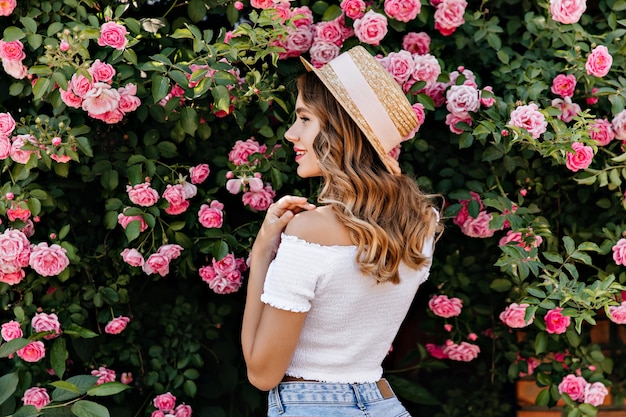 Retrato de trás de uma linda garota caucasiana, olhando para rosas cor de rosa. foto ao ar livre do elegante modelo feminino em pé de chapéu perto de arbusto em flor.