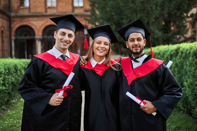 Retrato de três amigos de pós-graduação sorridentes com vestes de formatura no campus da universidade com diploma.