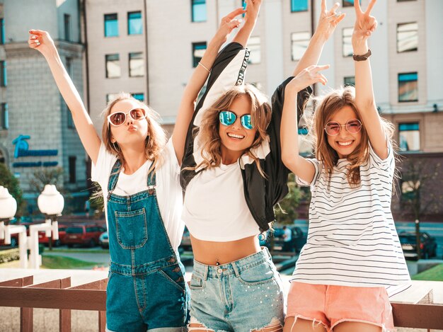 Retrato de três jovens bonitas garotas hipster sorridente em roupas da moda no verão. Mulheres despreocupadas sexy, posando na rua. Modelos positivos, se divertindo em óculos de sol.