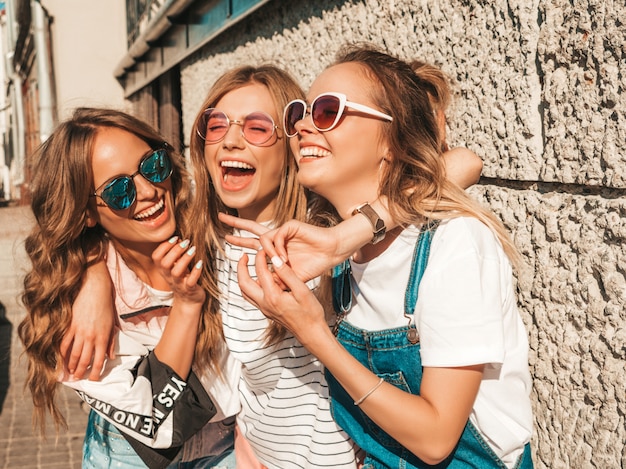 Retrato de três jovens bonitas garotas hipster sorridente em roupas da moda no verão. Mulheres despreocupadas sexy, posando na rua. Modelos positivos, se divertindo em óculos de sol.