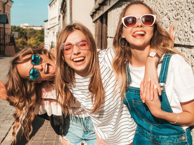 Retrato de três jovens bonitas garotas hipster sorridente em roupas da moda no verão. Mulheres despreocupadas sexy posando perto da parede na rua. Modelos positivos se divertindo em óculos de sol