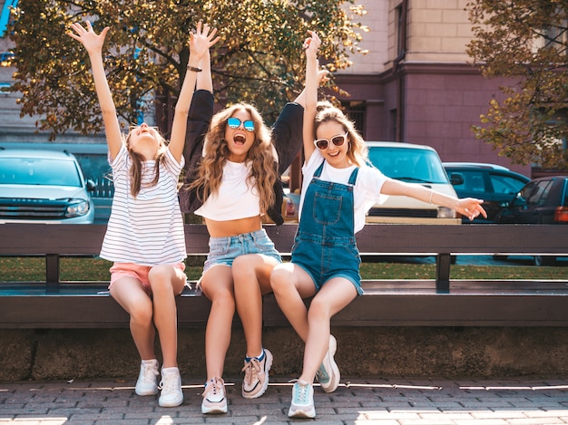 Retrato de três jovens bonitas hipster garotas sorridentes em roupas da moda no verão. mulheres despreocupadas sexy, sentado no banco na rua. modelos positivos se divertindo em óculos de sol.