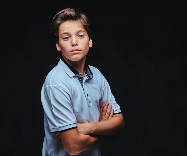 Retrato de um adolescente vestido com uma camiseta branca em pé com os braços cruzados. Isolado em um fundo escuro.