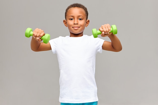 Retrato de um adorável garoto atlético de pele escura em uma camiseta branca em branco fazendo uma rotina de exercícios físicos matinais para bíceps usando dois halteres verdes com uma expressão facial alegre e energética