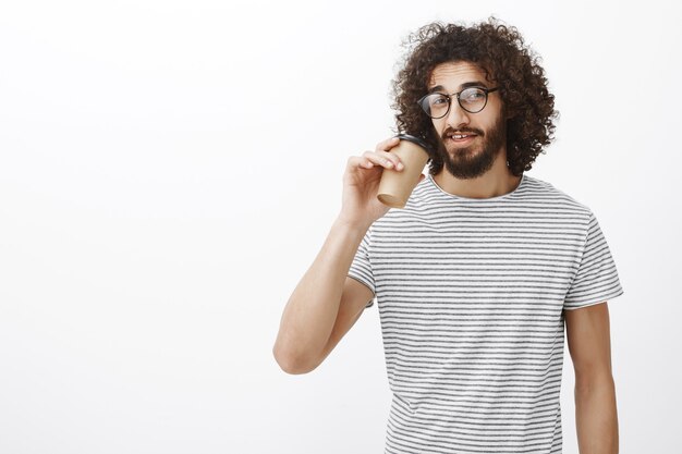 Retrato de um barista magro e confiante, com uma camiseta da moda e óculos, bebendo café em uma xícara e olhando