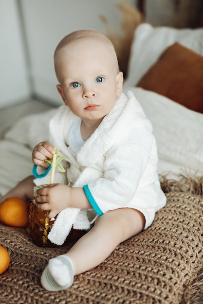 Foto grátis retrato de um bebezinho lindo com cabelo curto em pijama branca sentado na cama com suco de limão