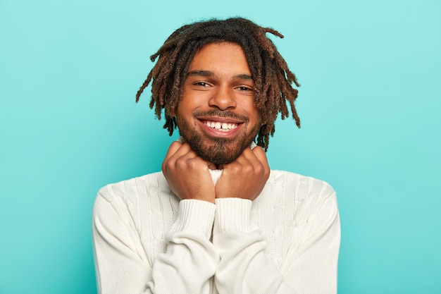 Retrato de um belo hipster alegre mantém as mãos embaixo do queixo, tem pele escura, dreadlocks, expressa emoções positivas, usa um macacão branco quente, está de bom humor, isolado sobre um fundo azul