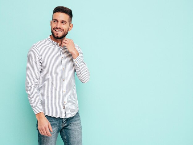 Retrato de um belo modelo sorridente Sexy homem elegante vestido de camisa e jeans Moda hipster masculino posando perto da parede azul no estúdio Alegre e feliz