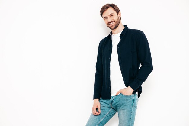 Retrato de um belo modelo sorridente Sexy homem elegante vestido de camisa e jeans Moda hipster masculino posando perto da parede branca no estúdio isolado