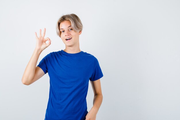 Retrato de um belo rapaz adolescente mostrando um gesto de aprovação em uma camiseta azul e uma alegre vista frontal