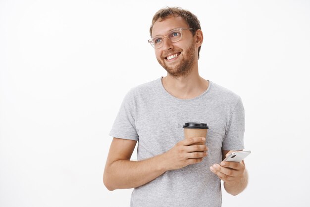 Retrato de um cara adulto caucasiano, alegre e satisfeito, com cabelo eriçado e loiro em copos, segurando um copo de papel com bebida e smartphone olhando para a esquerda com um sorriso otimista e sonhador