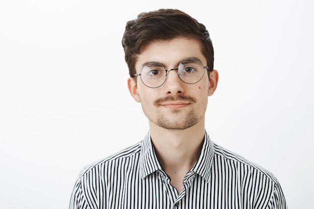 Retrato de um cara barbudo caucasiano confiante e amigável com bigode, usando óculos, sorrindo educadamente e parecendo relaxado e calmo sobre a parede cinza, ouvindo a reclamação do cliente sobre a parede cinza