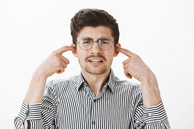 Foto grátis retrato de um cara comum choramingando chateado de óculos redondos e camisa listrada, cobrindo as orelhas com o dedo indicador, fazendo uma expressão de desagrado, sentindo antipatia ao ouvir um arranhão horrível no quadro-negro