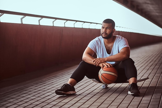 Retrato de um cara de pele escura pensativo, vestido com uma camisa branca e shorts esportivos, segura uma bola de basquete enquanto está sentado em um skate em uma passarela sob uma ponte.