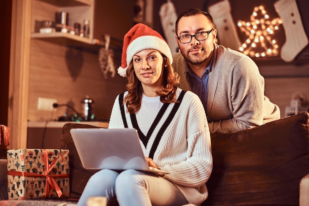 Retrato de um casal adorável com laptop olhando para a câmera comemorando a véspera de Natal em casa.