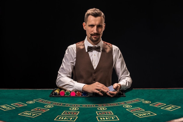 Retrato de um crupiê está segurando cartas de baralho, fichas de jogo na mesa. Fundo preto. Um jovem crupiê de camisa, colete e gravata borboleta está esperando por você na mesa de blackjack