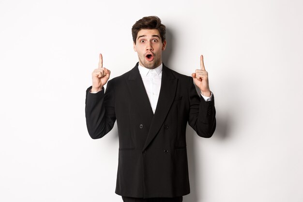 Retrato de um empresário bonito surpreso em um terno preto, dizendo uau e parecendo surpreso, apontando o dedo para o espaço da cópia, em pé sobre um fundo branco