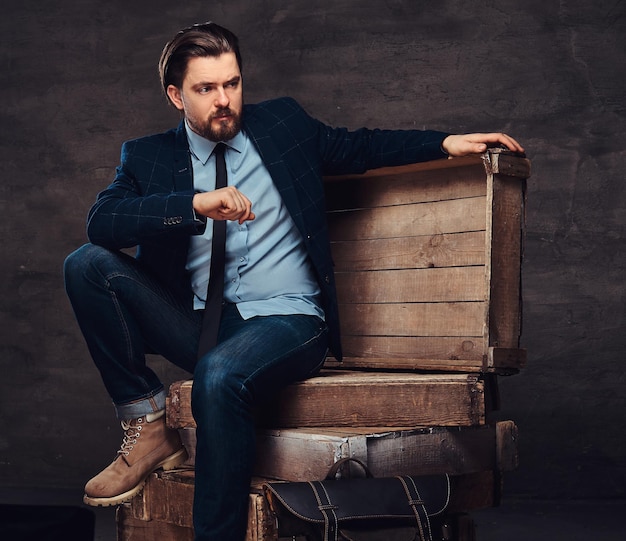 Retrato de um empresário de meia idade com cabelo elegante e barba, vestido de jeans, paletó e gravata, sentado em caixas de madeira em um estúdio. Isolado em um plano de fundo texturizado escuro.