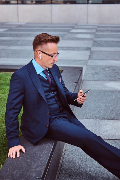 Retrato de um empresário elegante confiante vestido com um terno elegante usando um smartphone enquanto está sentado ao ar livre contra um fundo de arranha-céu.