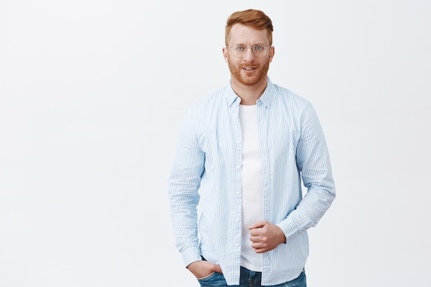 Foto grátis retrato de um empresário ruivo bem-sucedido, bonito e autoconfiante, de camisa e óculos, segurando o colarinho e sorrindo com uma expressão charmosa e sedutora sobre a parede cinza