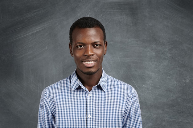 Foto grátis retrato de um estudante atraente de pele escura, vestindo uma camisa xadrez com uma expressão confiante e alegre, em pé na parede do quadro